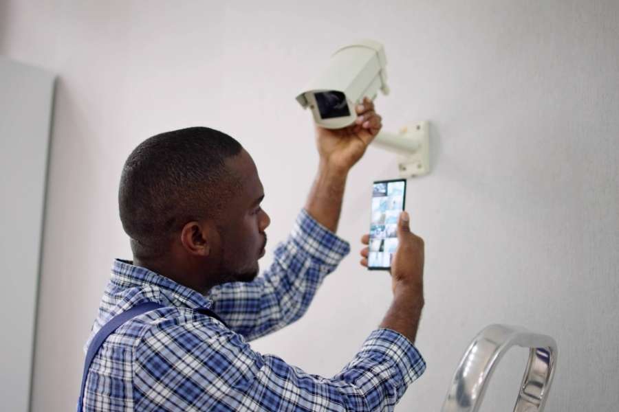 Un installateur professionnel ajuste une caméra de sécurité blanche tout en consultant son smartphone pour vérifier les réglages de l'équipement lié à notre Ajax systems avis.