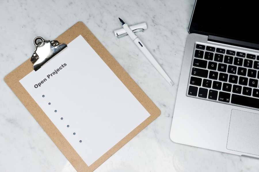 Un porte-bloc avec une feuille intitulée Open Projects posé à côté d'un stylo blanc et d'un ordinateur portable sur du marbre.