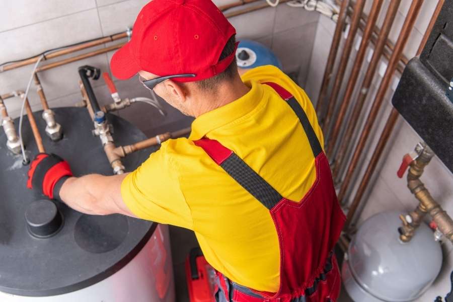 Un plombier-chauffagiste en combinaison rouge et jaune effectue la maintenance d'une chaudière complexe, une intervention qui sera détaillée sur la facture.