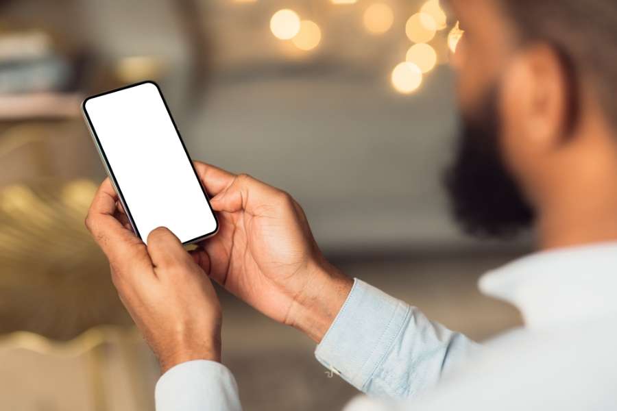 Un homme barbu, vu de dos, tient un smartphone avec un écran blanc vide, symbolisant sa relation aux écrans dans un intérieur chaleureux.