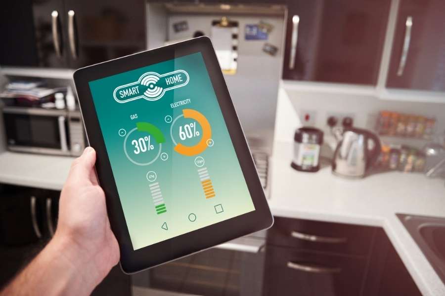Une tablette tactile affichant une application de maison connectée avec la consommation de gaz et d'électricité est tenue dans une cuisine moderne.